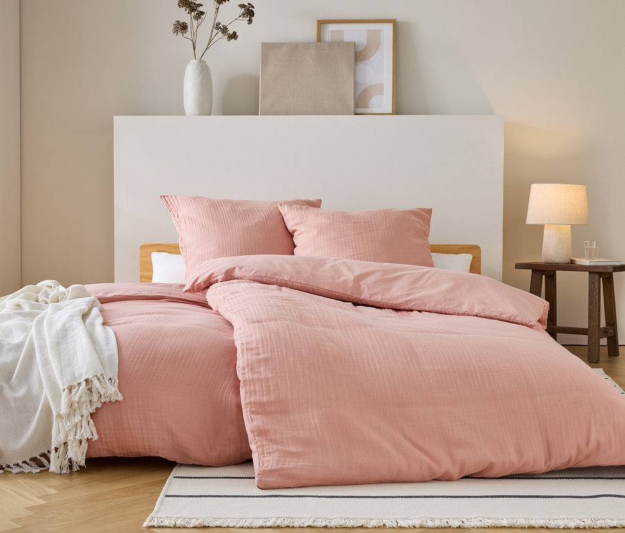 Ein Bett mit rosa Bettwäsche und Kissen steht auf einem gestreiften Teppich. Eine weiße Decke liegt auf dem Bett. Ein Nachttisch steht daneben.