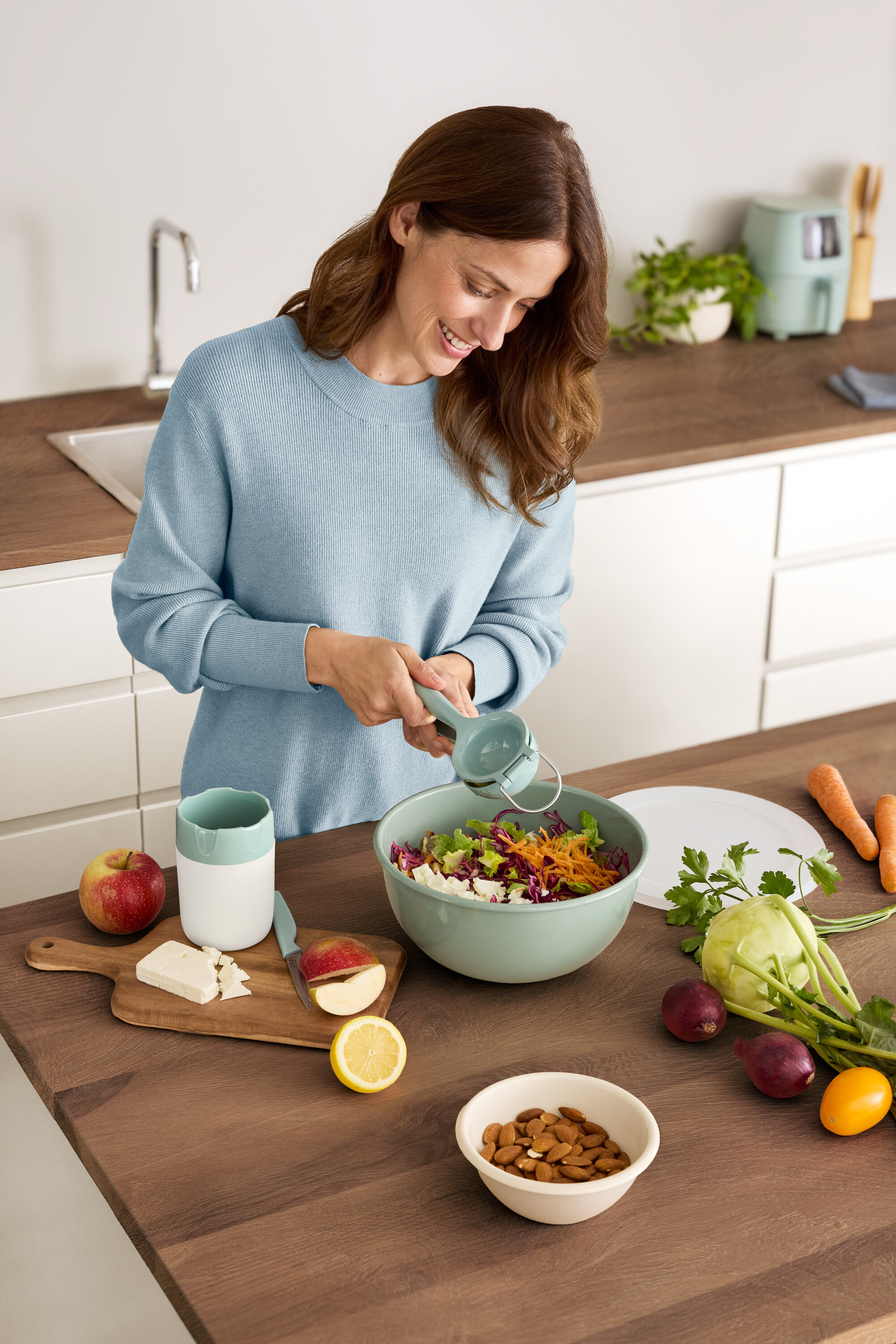 Frau presst Zitrone in eine Salatschüssel. Auf dem Tisch liegen ein Apfel, Käse, Mandeln und Gemüse.