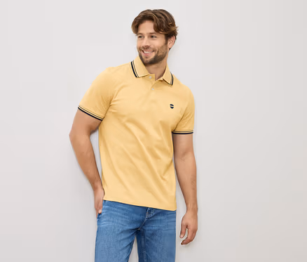 Mann mit gelbem Poloshirt und Jeans, stehend vor einer hellen Wand.