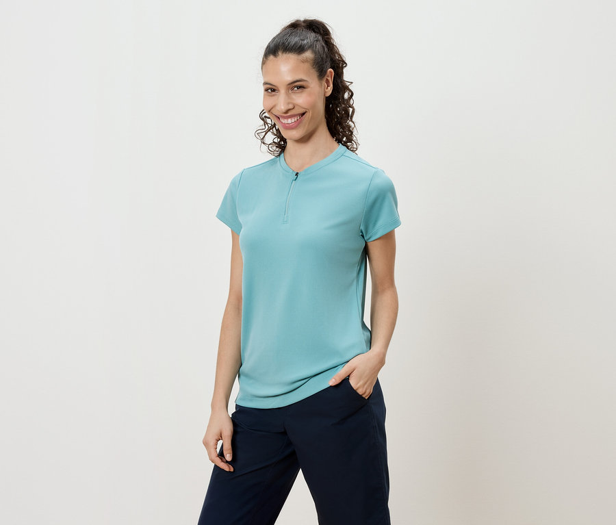 Frau mit lockigem Haar und Pferdeschwanz, die ein türkisfarbenes Shirt und marineblaue Hosen trägt.