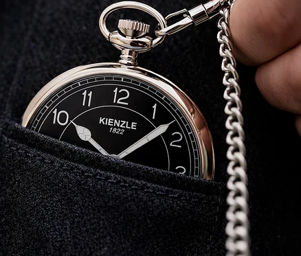 Nahaufnahme einer Person, die eine silberne Taschenuhr mit schwarzem Zifferblatt und Kette in einer dunklen Stofftasche hält.