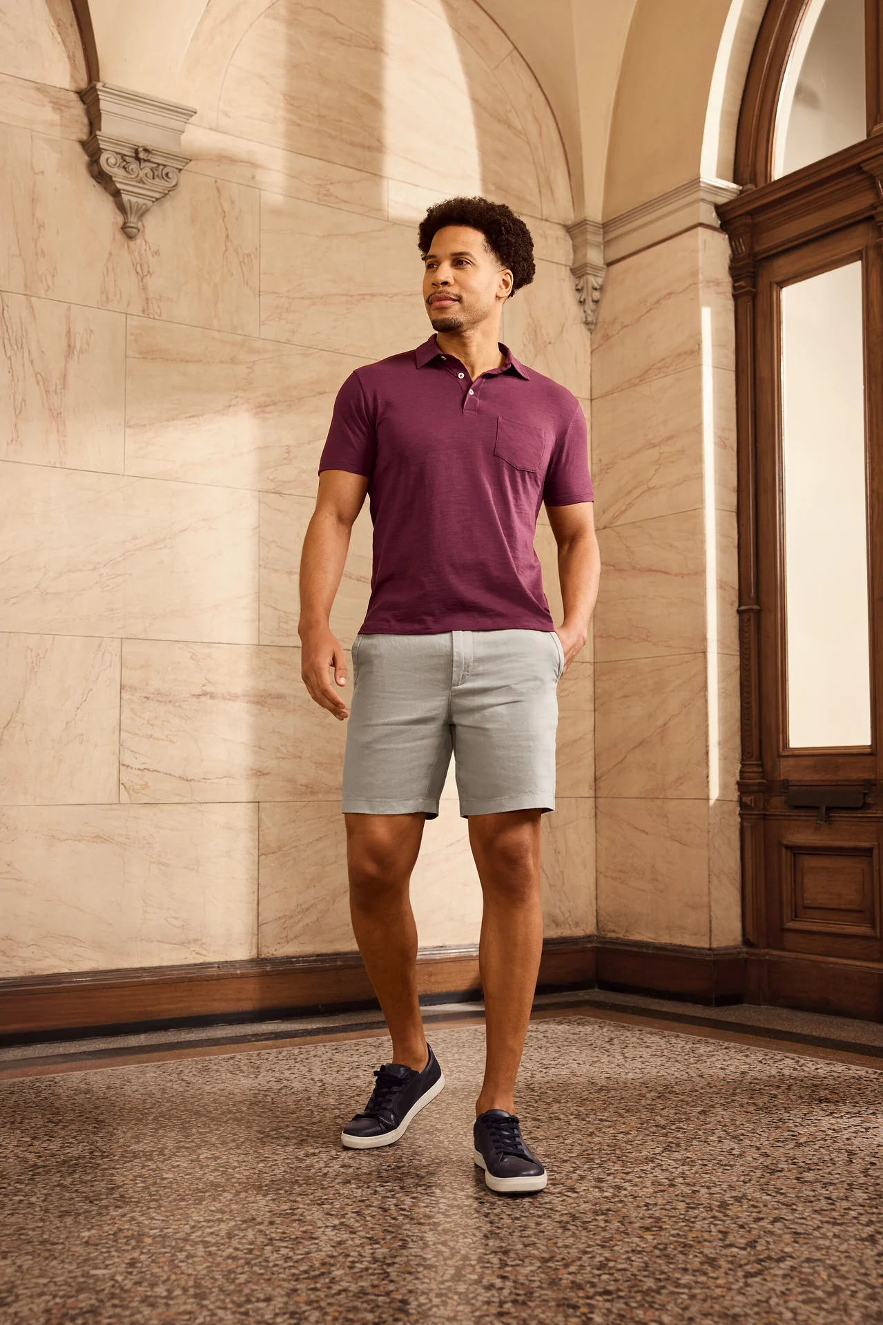 Mann mit lockigem Haar trägt weinrotes Poloshirt, Shorts und marineblaue Schuhe. Er steht vor einer verzierten Wand und einer Holztür.