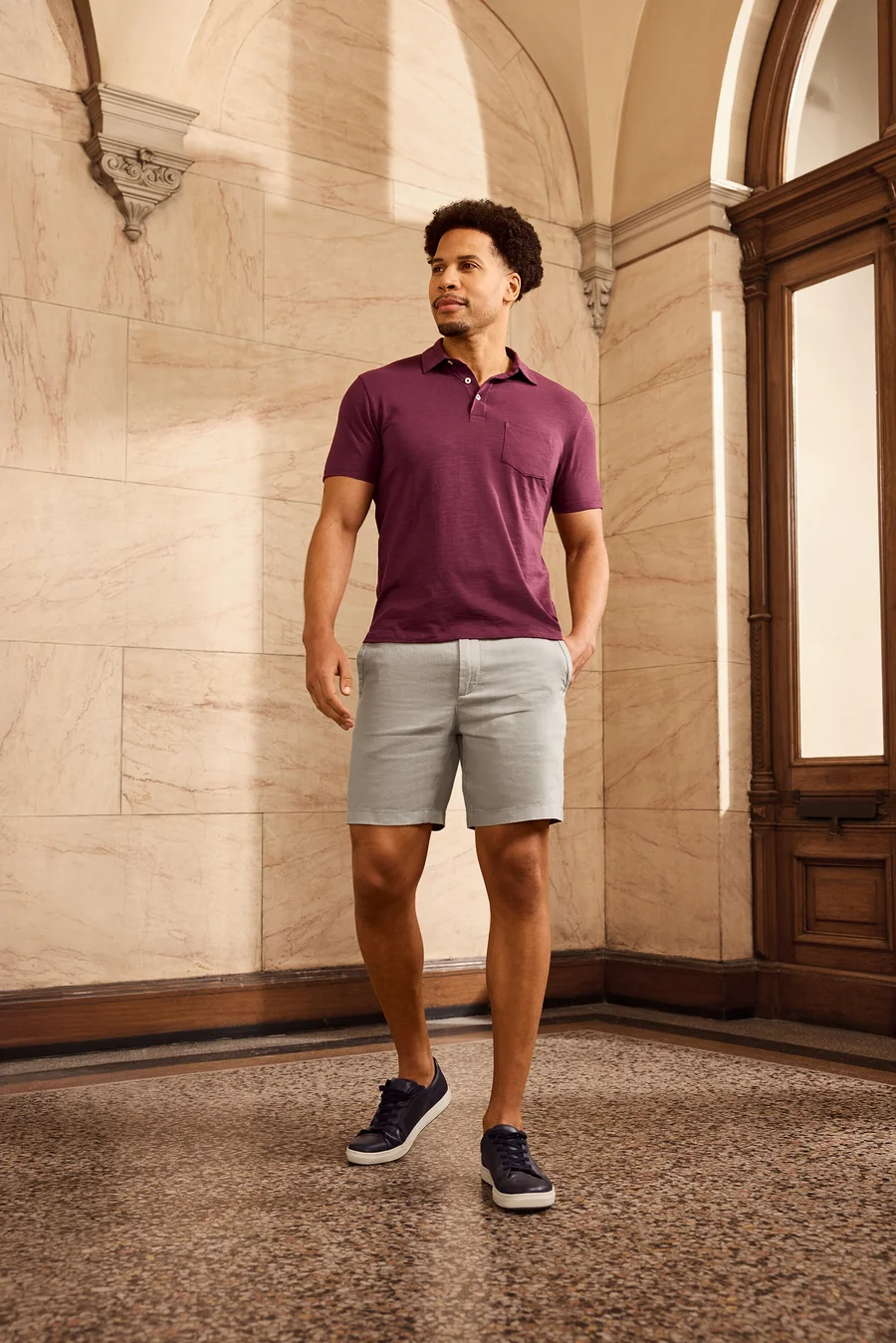 Mann mit lockigem Haar trägt weinrotes Poloshirt, Shorts und marineblaue Schuhe. Er steht vor einer verzierten Wand und einer Holztür.