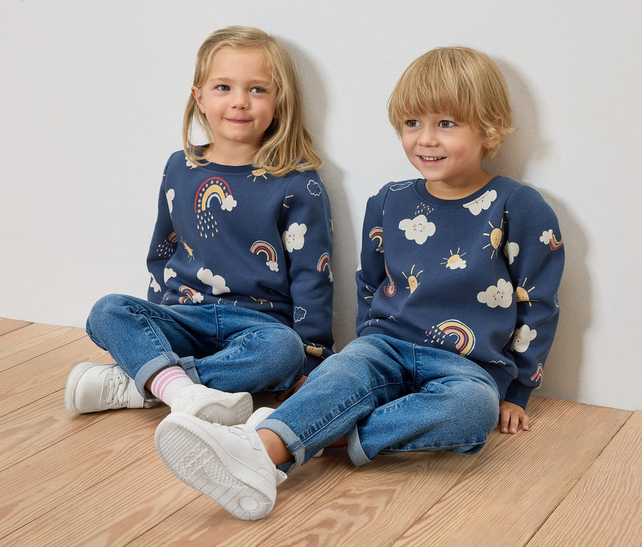 Zwei Kinder sitzen auf einem Holzboden vor einer Wand. Beide tragen dunkelblaue Kinder-Sweatshirts mit angerauter Innenseite, Jeans und Turnschuhe.
