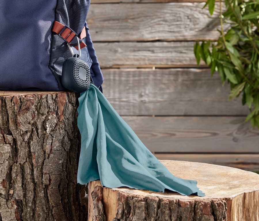 Ein marineblauer Rucksack liegt auf einem Baumstumpf, daran befestigt ist ein grauer Lautsprecher mit einem türkisfarbenen Tuch.