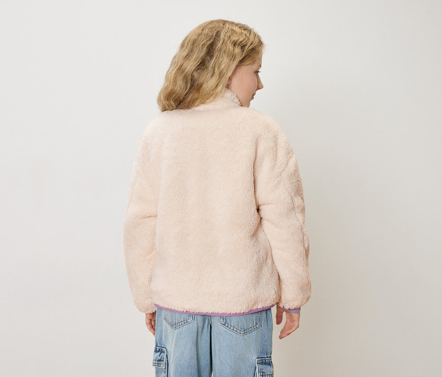 Rückansicht einer jungen Frau mit blonden, gewellten Haaren, die eine hellrosa Teddyfelljacke und blaue Jeans trägt.