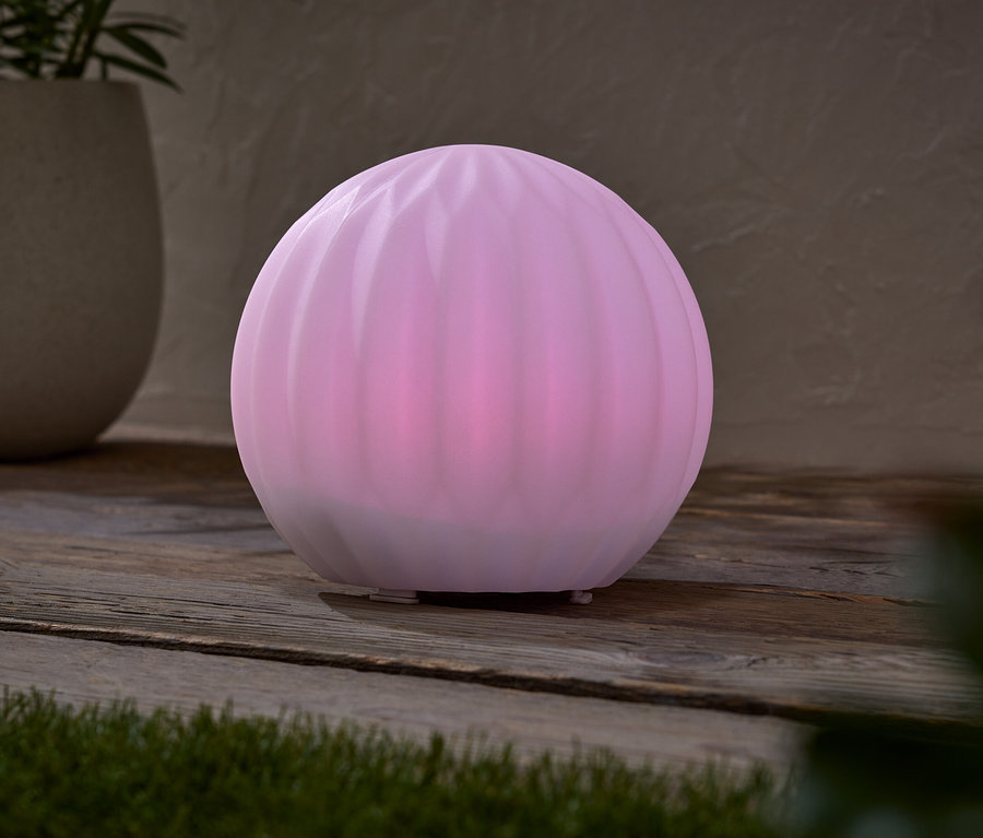 Eine rosafarbene Kugel-Lampe steht auf einem Holztisch, neben einem Topf mit einer Pflanze.