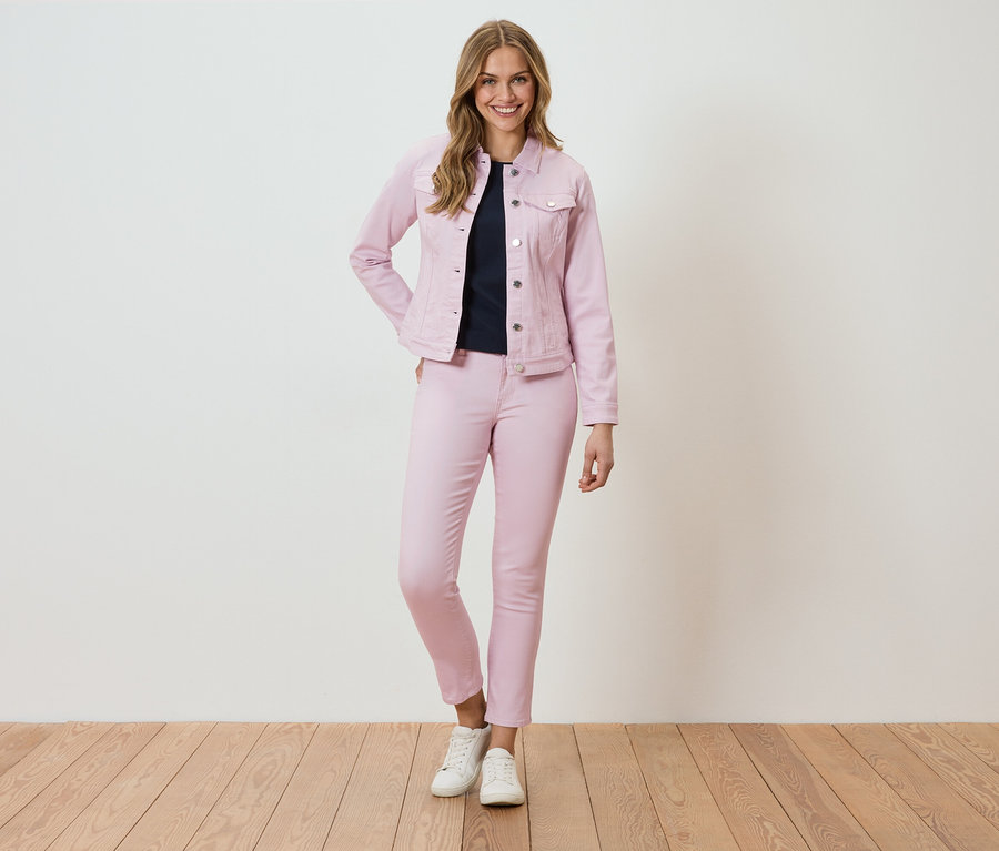 Frau steht vor einer Wand und trägt eine rosa Jeansjacke und Hose.