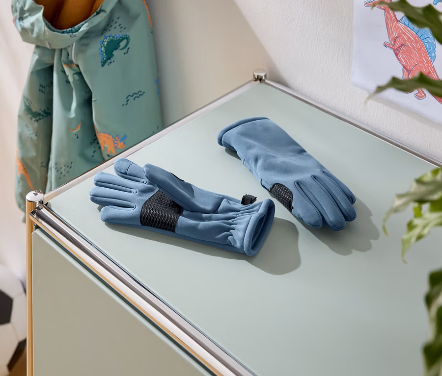 Auf einem Tisch liegen zwei blaue Handschuhe mit schwarzen Handflächen. Im Hintergrund sind eine Jacke mit Dinosaurier-Muster und ein Fußball zu sehen.