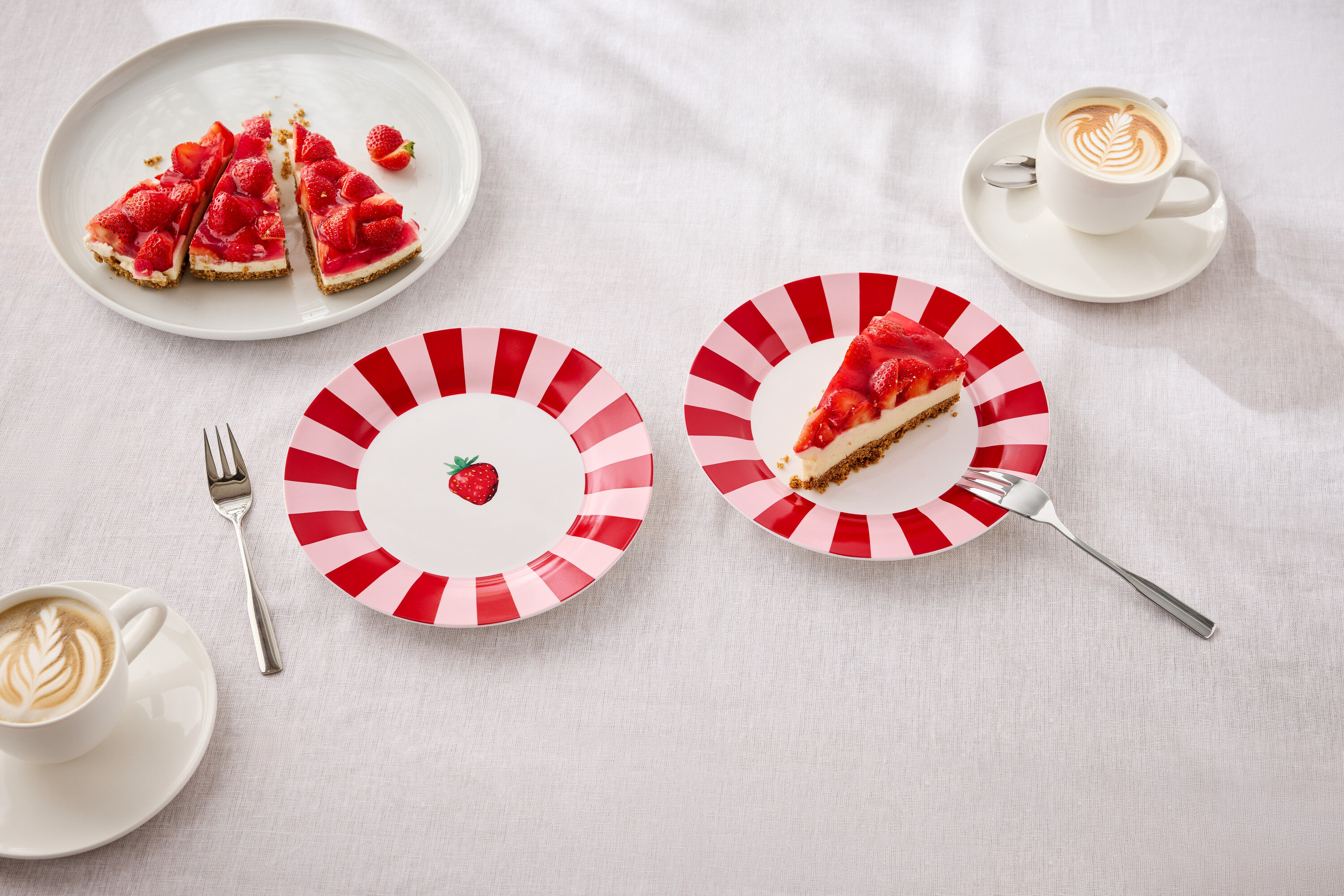 Auf einem Tisch liegen Teller mit Erdbeerkuchen, Gabeln und Tassen mit Latte Art.