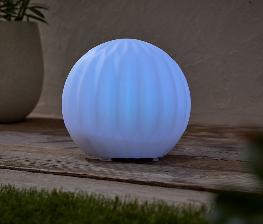 Eine leuchtende, kugelförmige Lampe steht auf einem Holztisch, neben einem Topf mit einer Pflanze.