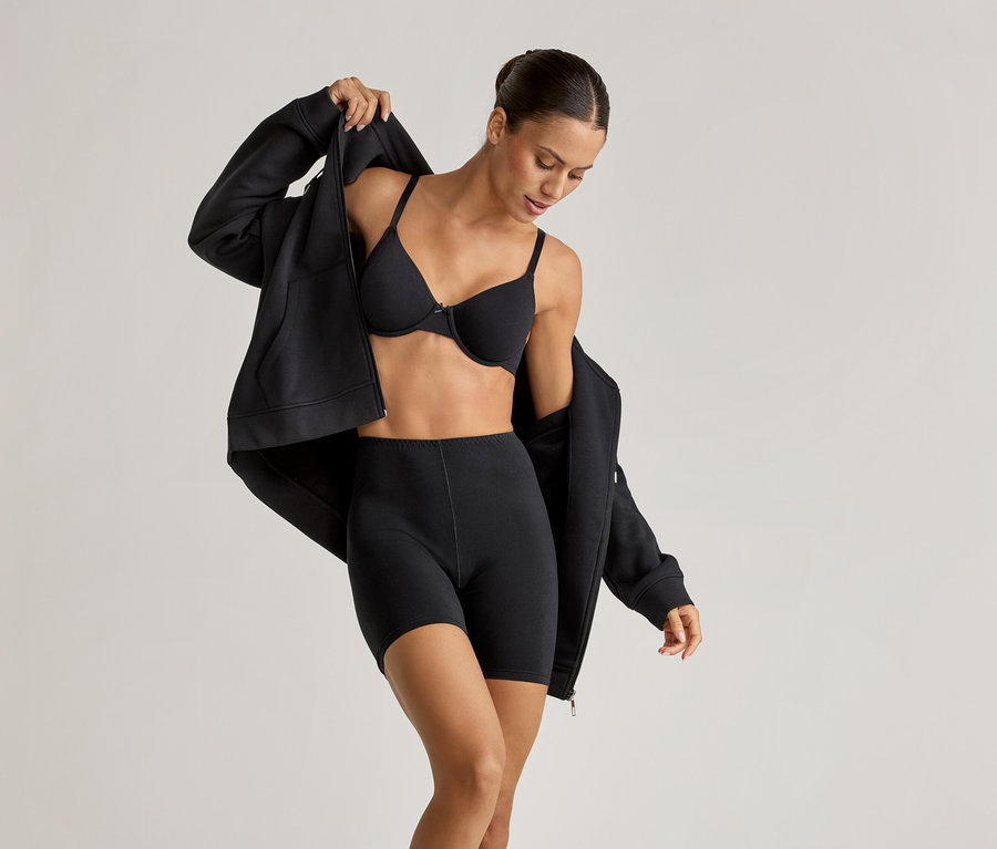 Frau mit schwarzem Bügel-BH, 2 Radler-Shorts und Lounge-Jacke, die sie öffnet.