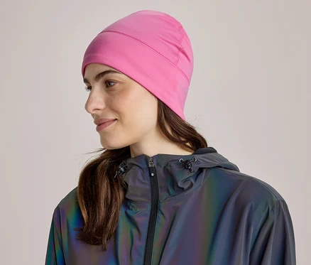 Frau mit braunen Haaren, die eine pinke Mütze und eine dunkle, reflektierende Jacke trägt, blickt nach rechts und lächelt leicht.
