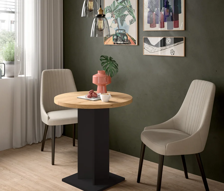 Ein kleiner Essbereich mit einem runden Holztisch, zwei beigefarbenen Polsterstühlen und einer dunkelgrünen Wand. Auf dem Tisch stehen eine rosa Vase, eine weiße Tasse und Macarons. Drei gerahmte Bilder und zwei Glas-Pendelleuchten hängen an der Wand, links ist ein Fenster.
