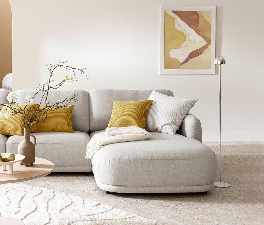Helles L-förmiges Sofa mit gelben und gestreiften Kissen, einer weißen Decke und einem runden Couchtisch mit Vase und Obstschale. Daneben steht eine silberfarbene SCHÖNER WOHNEN-Kollektion Akku-Stehleuchte »Bellis« vor einem abstrakten Wandbild.