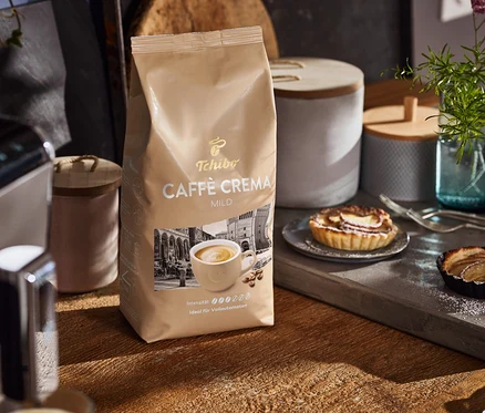 Auf einem Holztisch stehen eine Packung Caffè Crema Mild, eine Kaffeemaschine und Teller mit Kuchen.