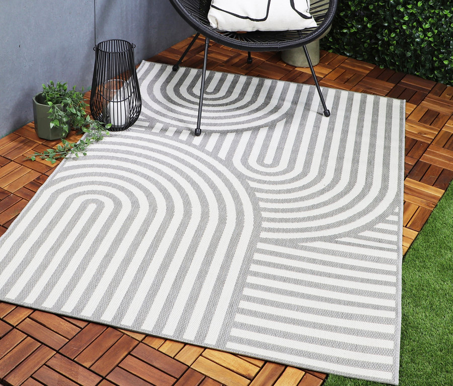 Outdoor-Teppich »Arc Symphony« mit weiß-grauem geometrischem Muster aus Bögen und Linien auf einem Holzdeck, neben einem schwarzen Stuhl, einer Laterne und grünen Pflanzen.