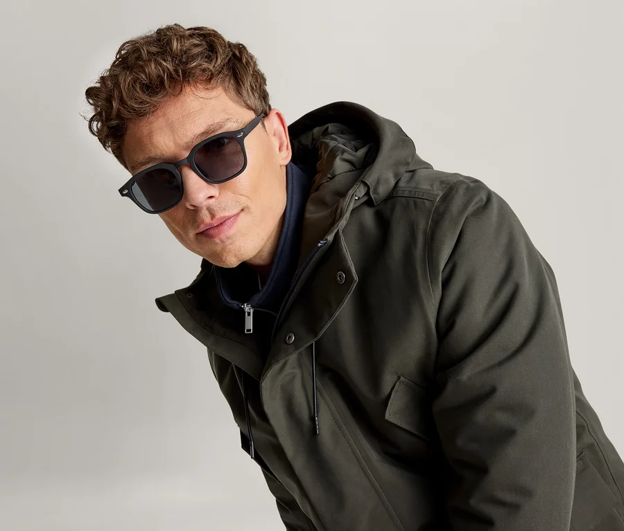 Mann mit lockigem braunem Haar, dunkler Sonnenbrille und dunkelgrünem Kapuzenparka, blickt in die Kamera.