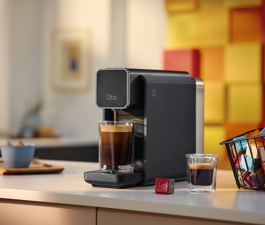 Eine schwarze Qbo Kaffeemaschine brüht Kaffee in ein doppelwandiges Glas auf einer weißen Arbeitsplatte. Daneben stehen ein weiteres Glas Kaffee, eine rote Kapsel und ein schwarzer Drahtkorb voller bunter Kaffeekapseln. Im Hintergrund sind eine blaue Schale mit Zuckerwürfeln auf einem Holztablett und eine Wand mit gelb-orangen Quadratmustern zu sehen.