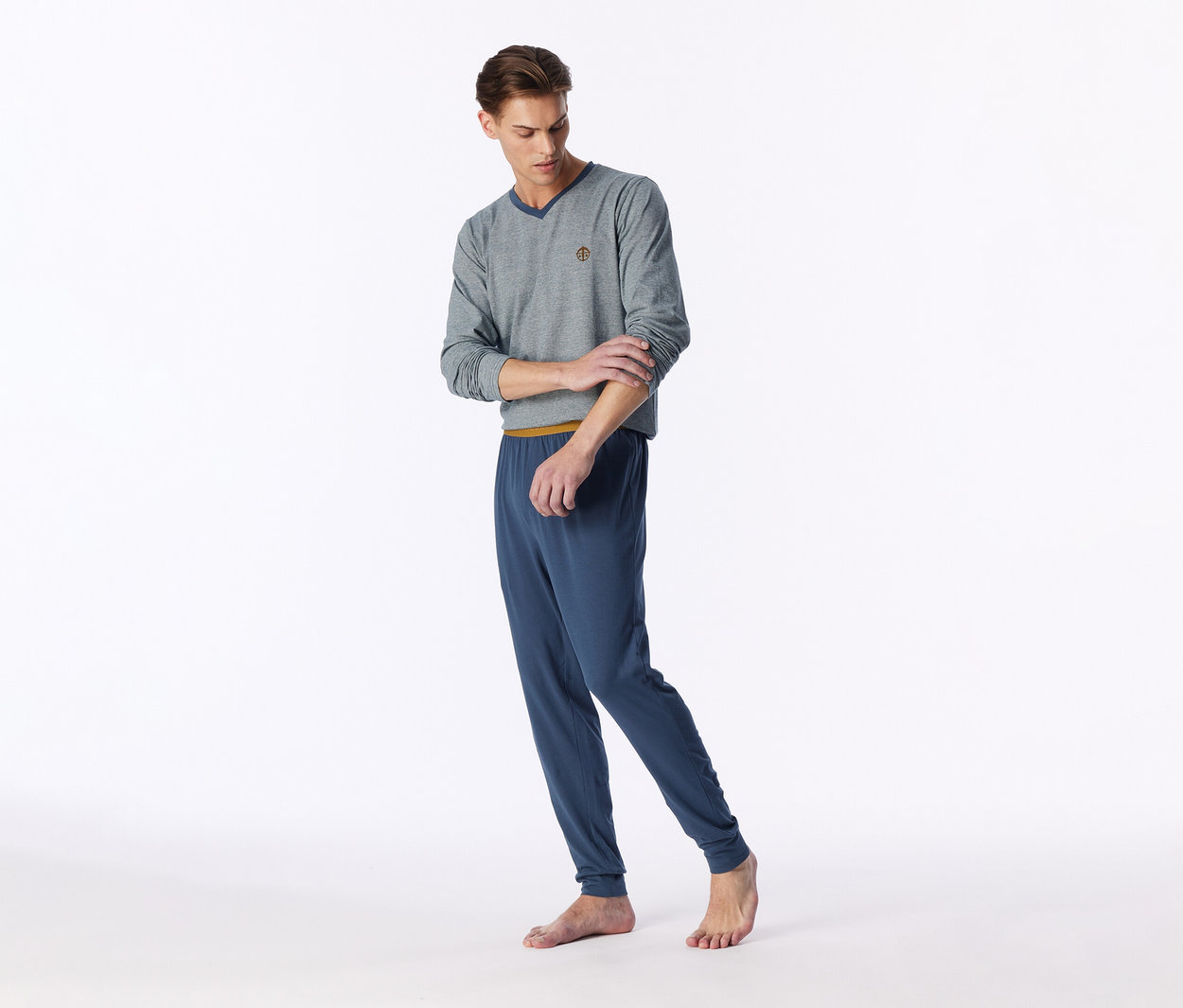 Junger Mann in grauem Langarmshirt mit V-Ausschnitt und blauer Pyjamahose, der barfuß steht und seinen Ärmel hochkrempelt.