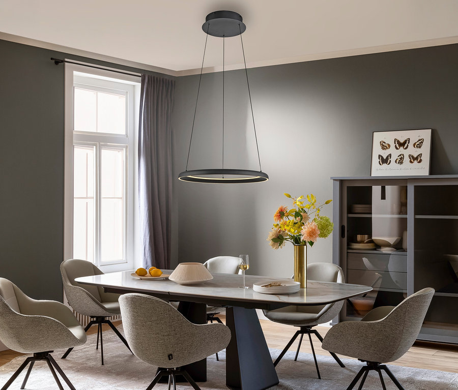 Ein modernes Esszimmer mit dunkelgrauen Wänden, einem rechteckigen Esstisch und sechs hellen Stühlen. Über dem Tisch hängt die beleuchtete schwarze Pendelleuchte »Quillo« der SCHÖNER WOHNEN-Kollektion. Der Tisch ist mit einer Vase bunter Blumen, Zitronen und Geschirr dekoriert. Rechts steht ein grauer Schrank mit Glastüren und einem Schmetterlingsbild, daneben ein großes Fenster mit grauen Vorhängen.
