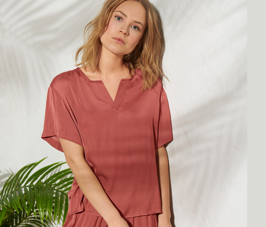 Frau posiert in Satin-Tunikabluse und Hose und hält einen Hut.