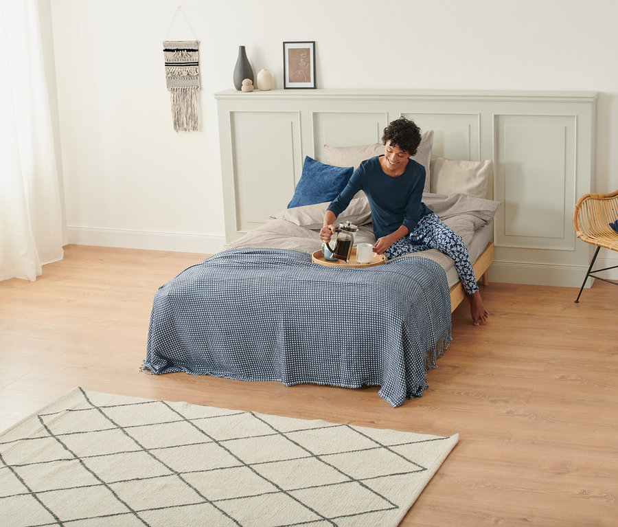 Frau im Pyjama gießt Kaffee aus einer French Press in eine Tasse auf einem Bett mit Wohndecke und Makramee-Wandteppich an der Wand.