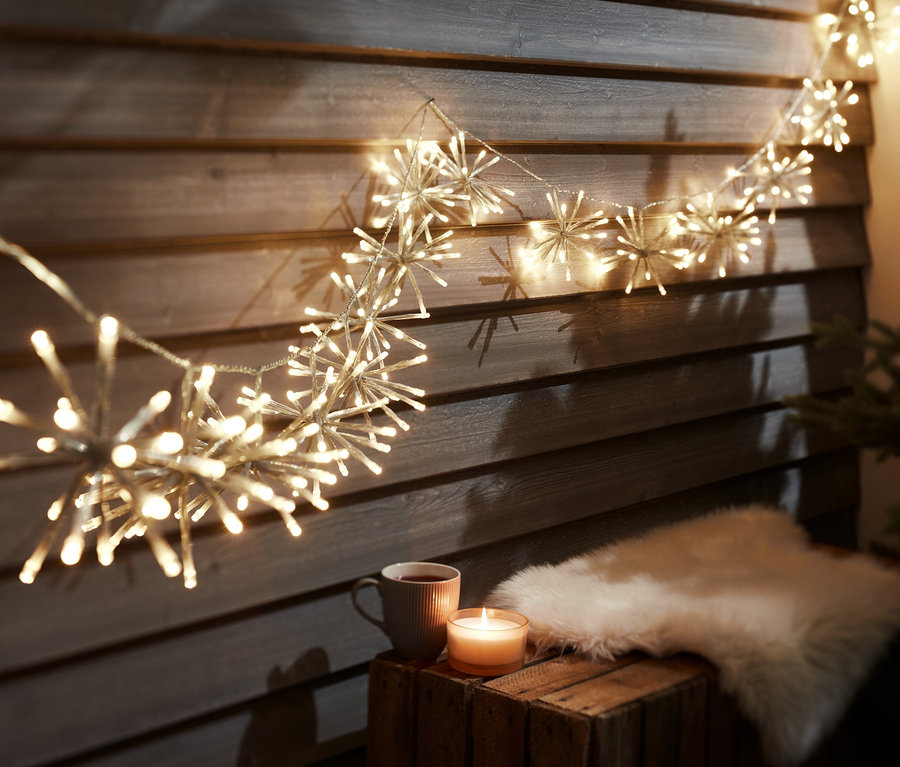 Eine LED-Outdoor-Lichtergirlande hängt an einer Holzwand über einer Tasse, einer Kerze und einem Fell.