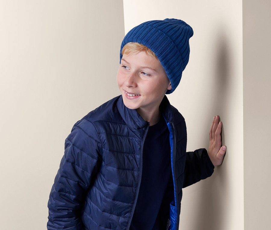Junge in blauer Steppjacke und Strickmütze lehnt an einer Wand.
