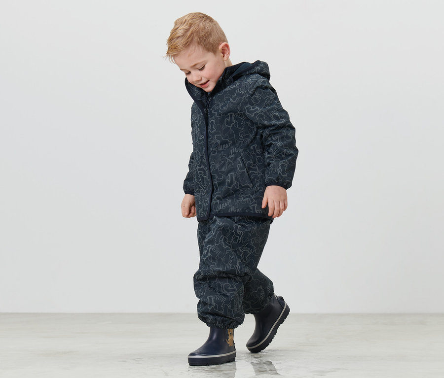 Junge trägt eine Kinder-Thermo-Regenjacke mit reflektierenden Elementen und dunkelblaue Gummistiefel und geht über den Boden.