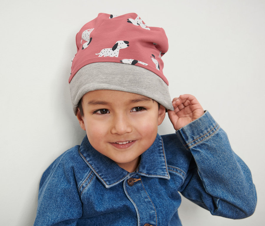 Ein Junge trägt eine rosa-graue Kinder-Wendemütze und eine Jeansjacke.
