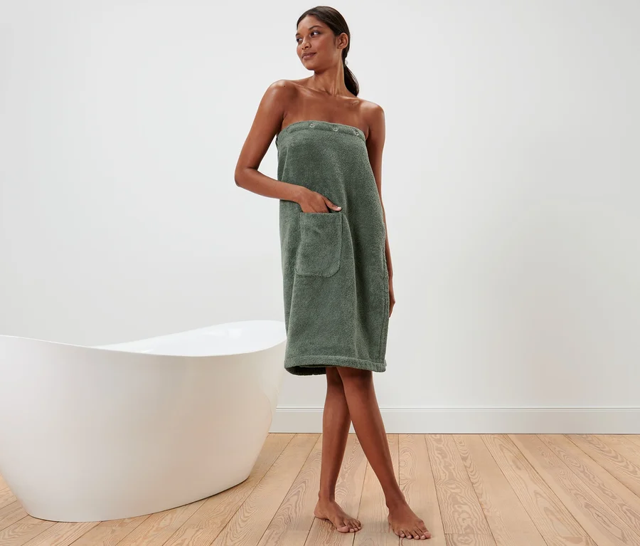 Frau trägt einen grünen Damen-Sauna-Kilt, neben einer weißen Badewanne stehend.