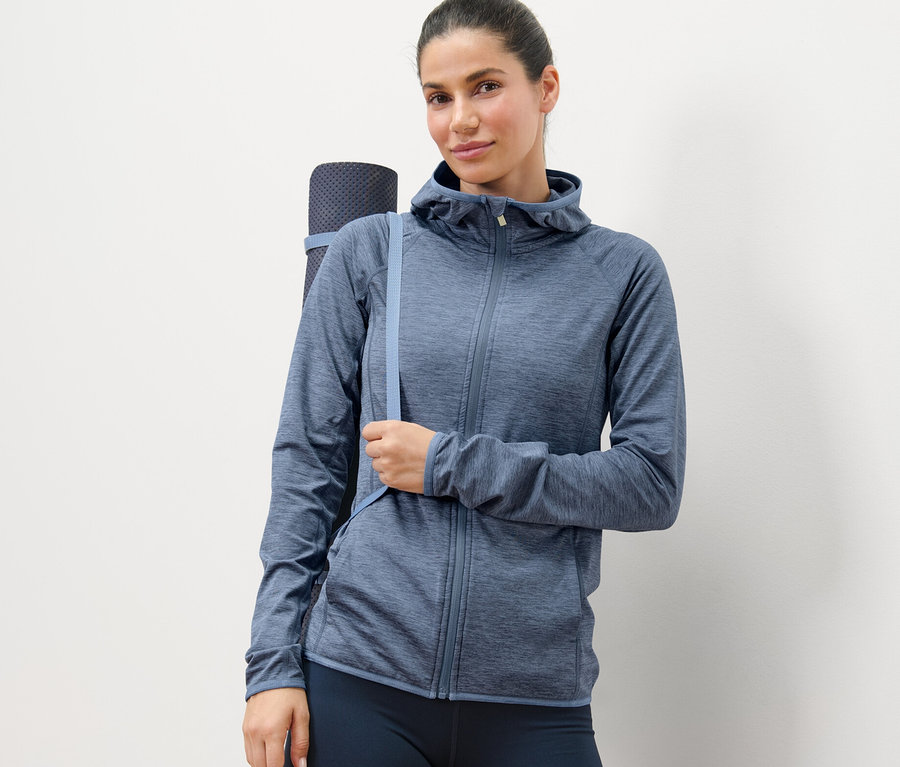 Frau posiert in einer blau melierten Sportjacke und trägt eine Yogamatte.