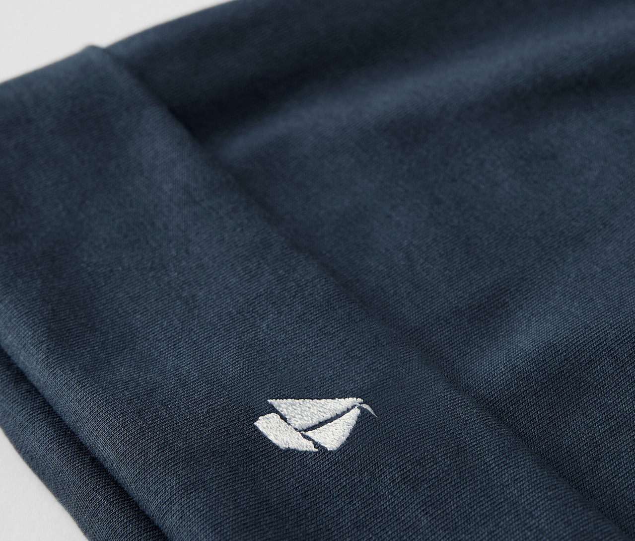 Detailaufnahme einer blauen Stoffmütze aus einem Baby-Set, mit einem aufgestickten weißen Logo.