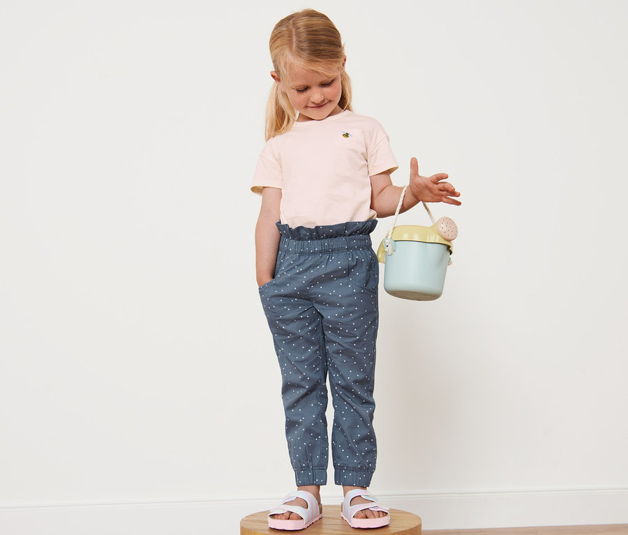 Ein Mädchen steht auf einem Holzsockel und hält eine blaugrüne Gießkanne. Sie trägt eine gepunktete Kleinkinder-Paperbaghose und rosa Sandalen.
