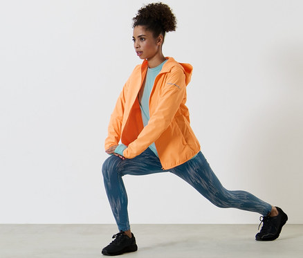 Frau dehnt sich in neonorangefarbener Windprotection-Laufjacke, hellblauem Langarm-Funktionsshirt und silberfarben reflektierender Sporttight.