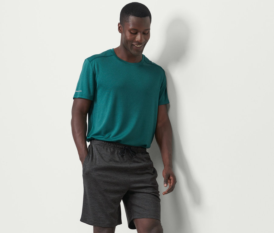 Mann in grünem T-Shirt und anthrazitfarbenen Sportshorts steht an einer weißen Wand.