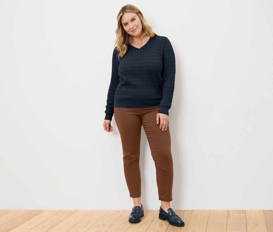 Eine Frau posiert in einem dunkelblauen Zopfstrickpullover und einer cognacfarbenen Stretchhose.