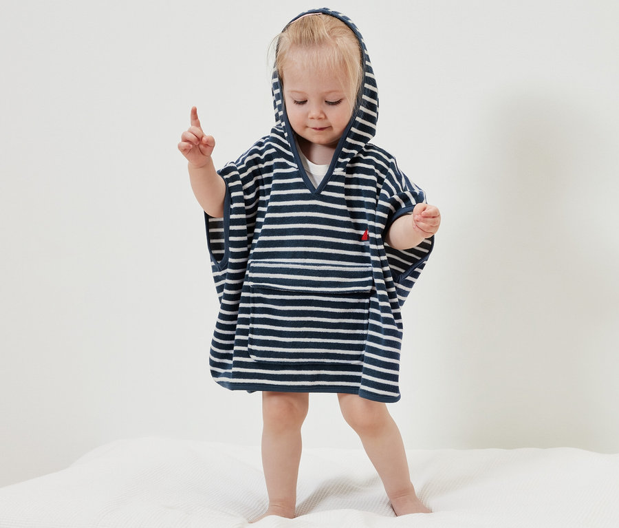Kleines Mädchen steht auf einer weißen Decke und trägt einen Frottierponcho im maritimen Stil.