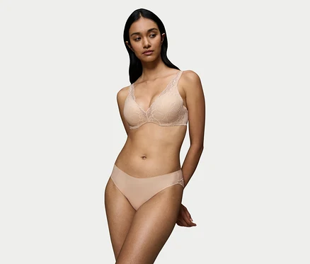 Eine Frau mit dunklem Haar trägt einen nude beigen Triumph Body Makeup Illusion Lace WP BH mit floralen Spitzendetails und passende Höschen. Ihre Hände sind hinter dem Rücken verschränkt.