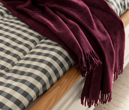 Pflaumenfarbene SCHÖNER WOHNEN-Kollektion Wohndecke »BASICS« mit Fransen liegt auf einem Bett mit grau-weiß karierter Bettwäsche.