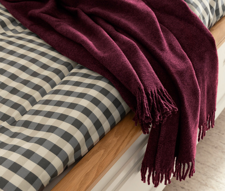 Eine weinrote Decke mit Fransen liegt auf einem Bett mit Karomuster.