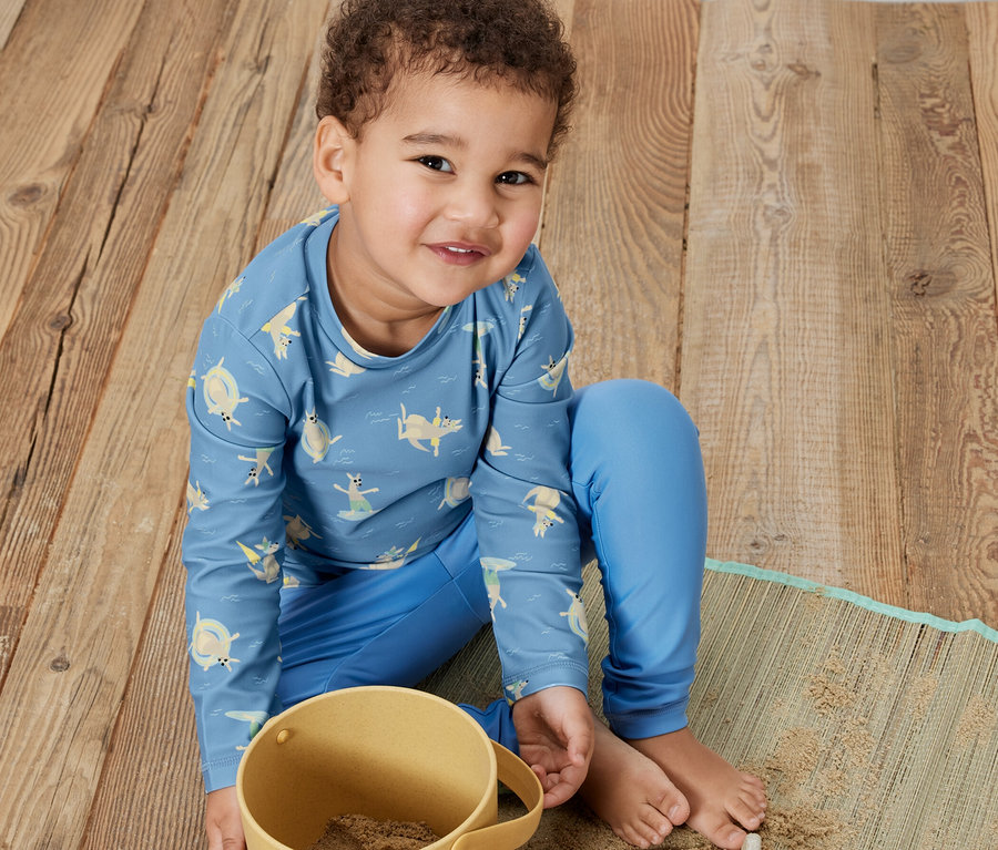 Ein Junge sitzt auf einem Holzboden und spielt mit Sand in einem gelben Eimer. Er trägt ein blaues Kinder-Shirt mit UV-Schutzfaktor 80, Känguru.