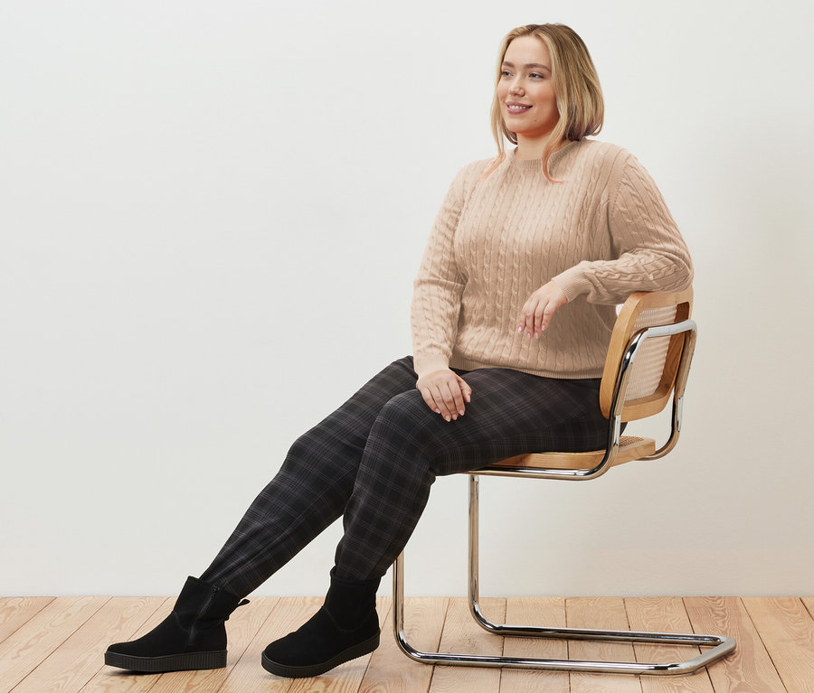 Frau sitzt auf einem Stuhl und trägt einen cremefarbenen Zopfstrickpullover und eine karierte Joggpants.