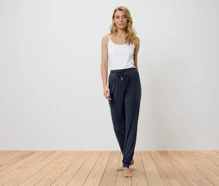 Blonde Frau steht auf einem Holzboden und trägt ein weißes Top und dunkelblaue Hosen.