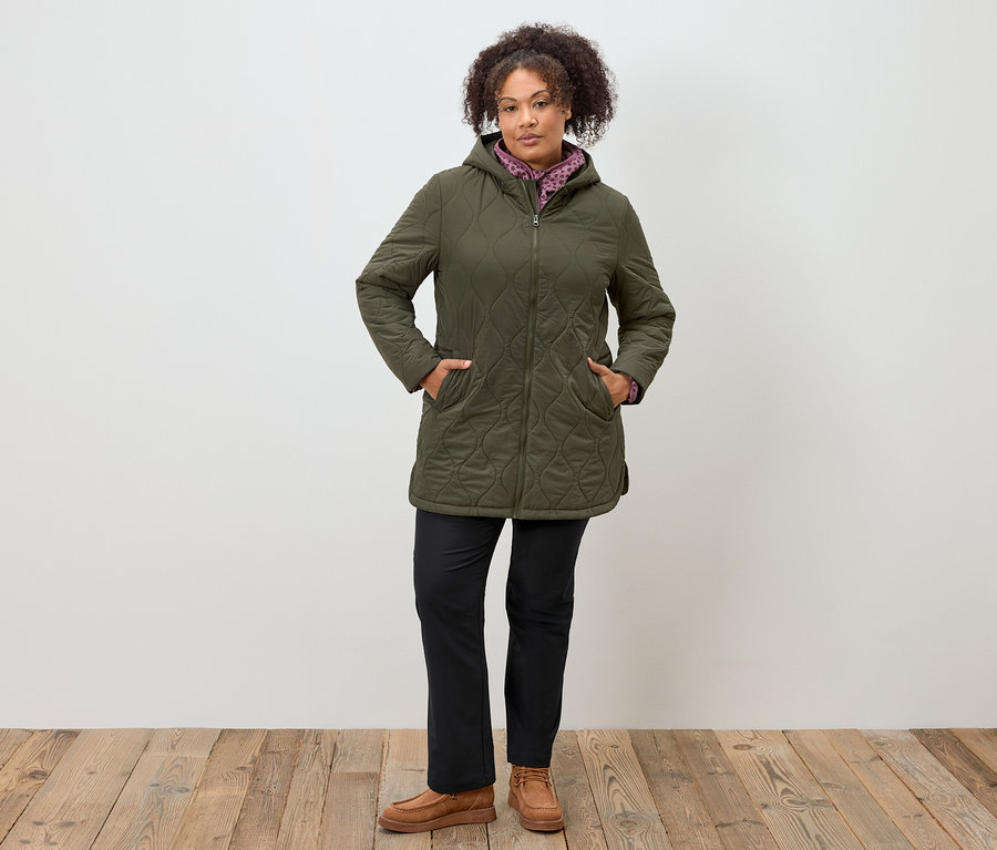 Frau steht mit Händen in den Taschen einer olivgrünen Steppjacke mit Kapuze, schwarzen Hosen und braunen Mokassins.