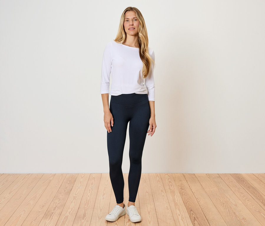 Eine Frau steht auf einem Holzboden und trägt ein weißes Langarmshirt und schwarze Leggings.