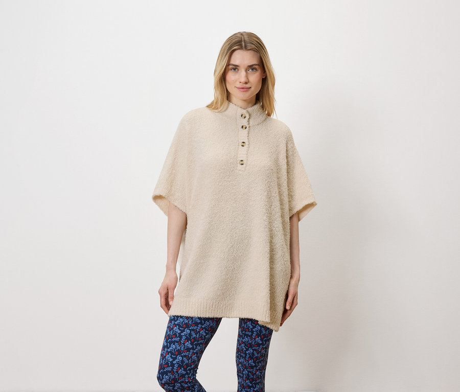 Eine Frau posiert im Studio vor einem weißen Hintergrund. Sie trägt einen beigen Pullover und blaue Leggings.
