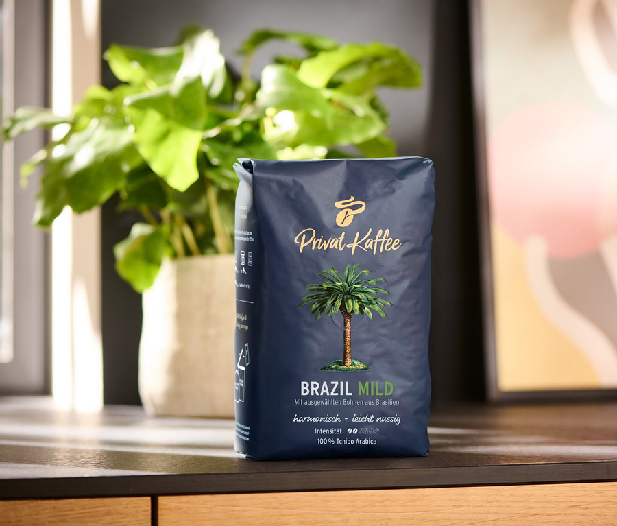 Eine 500g Packung Privat Kaffee Brazil Mild Ganze Bohne steht auf einer Holzoberfläche. Im Hintergrund befinden sich eine Pflanze im Topf und ein abstraktes Gemälde.