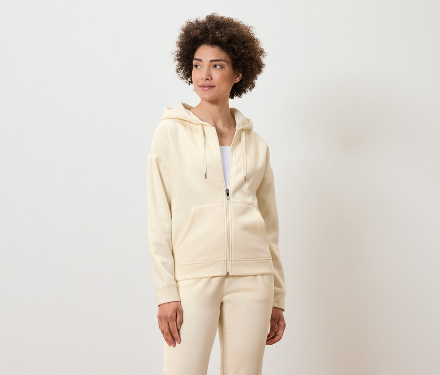 Frau mit lockigem Haar posiert in einem cremefarbenen Reißverschluss-Hoodie und Trainingshose.